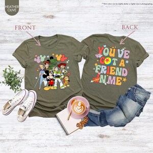 May include: Heather olive green t-shirts with a colorful graphic of Disney characters on the front and the text "YOU'VE GOT A FRIEND IN ME" on the back. The shirts are paired with jeans and white sneakers.