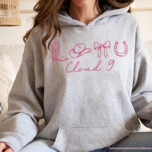 May include: A grey hoodie with a front pocket and hood. It displays a pink graphic of a cowboy boot, hat, bow, horseshoe, and the words "Cloud 9". The hoodie is labelled "Sport Grey".