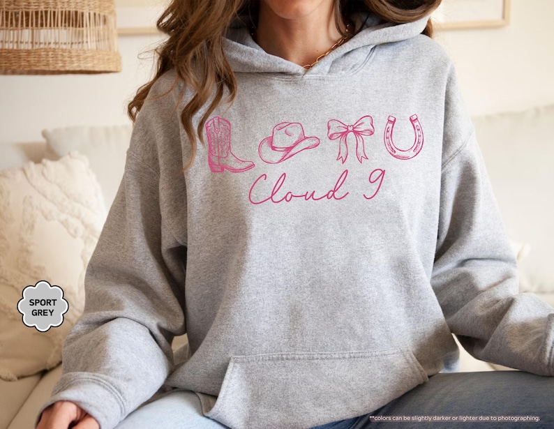 May include: A grey hoodie with a front pocket and hood. It displays a pink graphic of a cowboy boot, hat, bow, horseshoe, and the words "Cloud 9". The hoodie is labelled "Sport Grey".