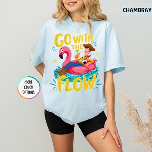 Puede incluir: Una camiseta azul claro con un gr&aacute;fico de un vaquero de dibujos animados sentado en un flotador de flamenco rosa. El texto dice GO WITH THE FLOW en letras amarillas. El dise&ntilde;o incluye salpicaduras de agua azules y estrellas amarillas. Color CHAMBRAY.