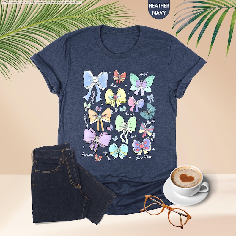 May include: Heather navy blue t-shirt with a colourful bow design and princess names. The bows are in various colours and styles, with names like Ariel, Cinderella, and Snow White. A pair of dark blue jeans and a cup of coffee are also in the image.
