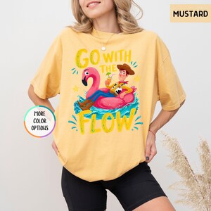 Puede incluir: Una camiseta color amarillo mostaza con un gr&aacute;fico de un vaquero sentado en un flotador de flamenco rosa sobre agua azul. El texto "GO WITH THE FLOW" aparece en letras amarillas. Una peque&ntilde;a insignia circular a la izquierda dice "MORE COLOR OPTIONS".