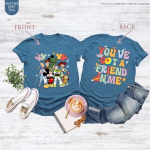 May include: Teal t-shirts with a cartoon graphic of Disney characters on the front and the phrase "YOU'VE GOT A FRIEND IN ME" on the back. The shirts are displayed with white sneakers and denim jeans.