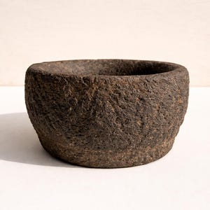 Puede incluir: Un cuenco de piedra marrón oscuro y texturizado con una parte superior ancha y abierta. El cuenco tiene forma redondeada y se asienta sobre una superficie blanca. El exterior del cuenco tiene una textura rugosa y desigual.