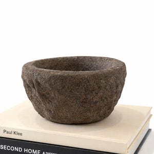 May include: A dark grey, roughly textured stone bowl. The bowl is round with a wide opening and rests on two books. The books have white and black covers, with the titles "Paul Klee" and "SECOND HOME A DIFFERENT WAY OF LIVING".