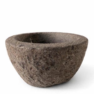 Puede incluir: Un mortero de piedra pequeño y redondo con una superficie rugosa y texturizada. El mortero es de color marrón apagado y tiene forma de cuenco ancho y poco profundo. El interior es oscuro y liso, contrastando con el exterior.