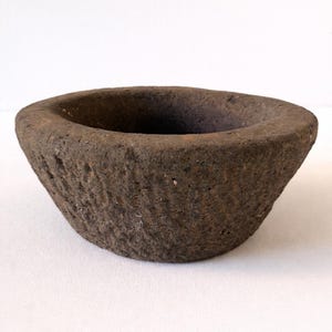 May include: A weathered, dark brown stone bowl with a textured surface. The bowl has a wide, shallow design, suitable for serving or display. The interior is a deep, dark colour.