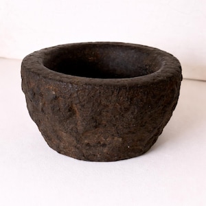 Puede incluir: Un mortero de piedra marrón oscuro, de textura rugosa, con un borde ancho y redondeado. El mortero tiene un interior profundo y un exterior ligeramente irregular, lo que sugiere un aspecto hecho a mano o envejecido. El cuenco está sobre una superficie blanca.