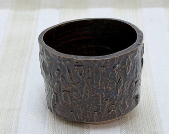 May include: A small, handcrafted ceramic bowl with a textured, bark-like exterior. The bowl is a dark, earthy brown color, with a smooth, dark interior. The cylindrical shape and unique texture give it a rustic, natural aesthetic.