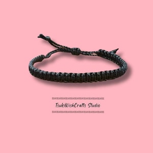 May include: Black macrame bracelet with adjustable knot closure. The bracelet is made of woven cord and is displayed on a pink background. The text "TsukiWishCrafts Studio" is below the bracelet.