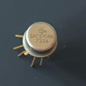 Op de afbeelding: Een klein, metalen elektronisch component met meerdere goudkleurige pinnen. Het component heeft een cirkelvormige bovenkant met de tekst "SFC2709A" en "7334" erop gedrukt, samen met een klein symbool. Waarschijnlijk gebruikt in elektronica.