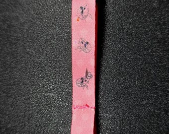 May include: A pink lanyard with a black plastic clasp and a silver-colored metal clip. The lanyard features a repeating pattern of small, black line drawings of rabbits.