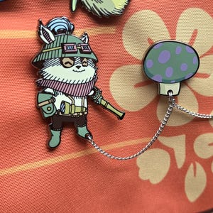 May include: Enamel pins featuring a cartoon character with goggles, a hat, and a scarf, holding a weapon. The pins are connected to two mushroom-shaped pins with a silver chain. The background is a floral pattern.
