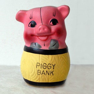 May include: A ceramic piggy bank featuring a pink pig head with black eyes and painted features, resting on a yellow barrel-shaped base with a textured surface. The words PIGGY BANK are printed in black on the front of the yellow barrel section.