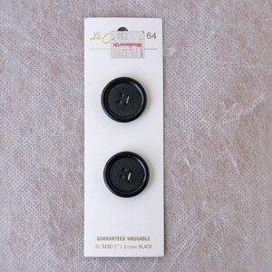 May include: Two black, round buttons on a white card. The card has text that reads "GUARANTEED WASHABLE 25 MM (1") 2 COUNT BLACK". The buttons are 1 inch in diameter and have four holes. The card also has a price tag.