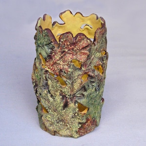 May include: A cylindrical ceramic candle holder featuring a textured exterior composed of overlapping oak leaf shapes in shades of green, brown, and yellow. The interior is finished in a smooth, solid yellow glaze, with cutouts between the leaves.