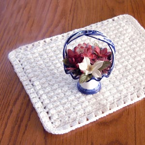 May include: A white crocheted placemat with a textured pattern. A small ceramic basket with a blue and white design holds red and white flowers. The basket has a handle and sits on the placemat.