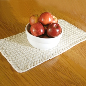 May include: A white ceramic bowl filled with several shiny red apples, resting on a rectangular, off-white crocheted mat with a textured, grid-like pattern and a simple stitched border, creating a functional centerpiece for a dining table.