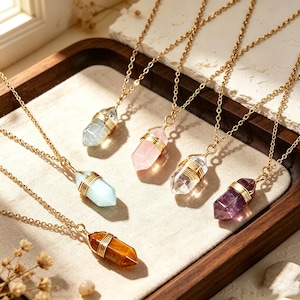 May include: A collection of six necklaces featuring raw, hexagonal crystal pendants in various colors including clear, pale pink, light blue, and deep purple, each wrapped in gold-toned wire and suspended from a delicate gold-toned metal chain.