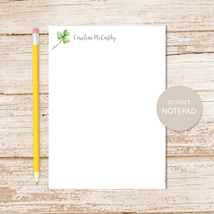 May include: A white notepad with a green shamrock and the name "Caroline McCarthy" printed at the top. The notepad is on a light wood background with a yellow pencil beside it. The notepad has a sticker that says "50 sheet notepad".