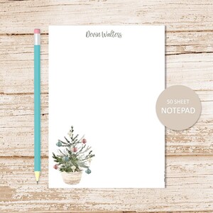 May include: A notepad with a watercolor illustration of a Christmas tree in a basket, adorned with ornaments. The notepad has the text "Devin Walters" at the top and a sticker that says "50 SHEET NOTEPAD". A light blue pencil is on the left.