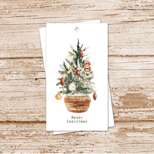 May include: Watercolor Christmas tree gift tags. The tags feature a decorated tree in a woven basket, adorned with ornaments and a nutcracker. The words "MERRY CHRISTMAS" are printed at the bottom. The tags are white with a hole at the top.