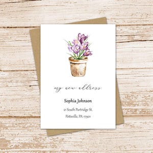 May include: A white rectangular card featuring a watercolor illustration of purple crocus flowers in a tan terracotta pot. The text reads: my new address, Sophia Johnson, 21 South Partridge St., Pottsville, PA 17901. The card sits on a brown envelope.