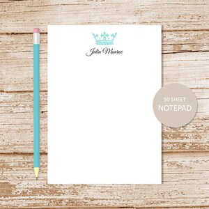 May include: A white notepad with a blue crown and the text "Julia Monroe" printed on the top. The notepad is on a wood background and has a label that says "50 sheet notepad".
