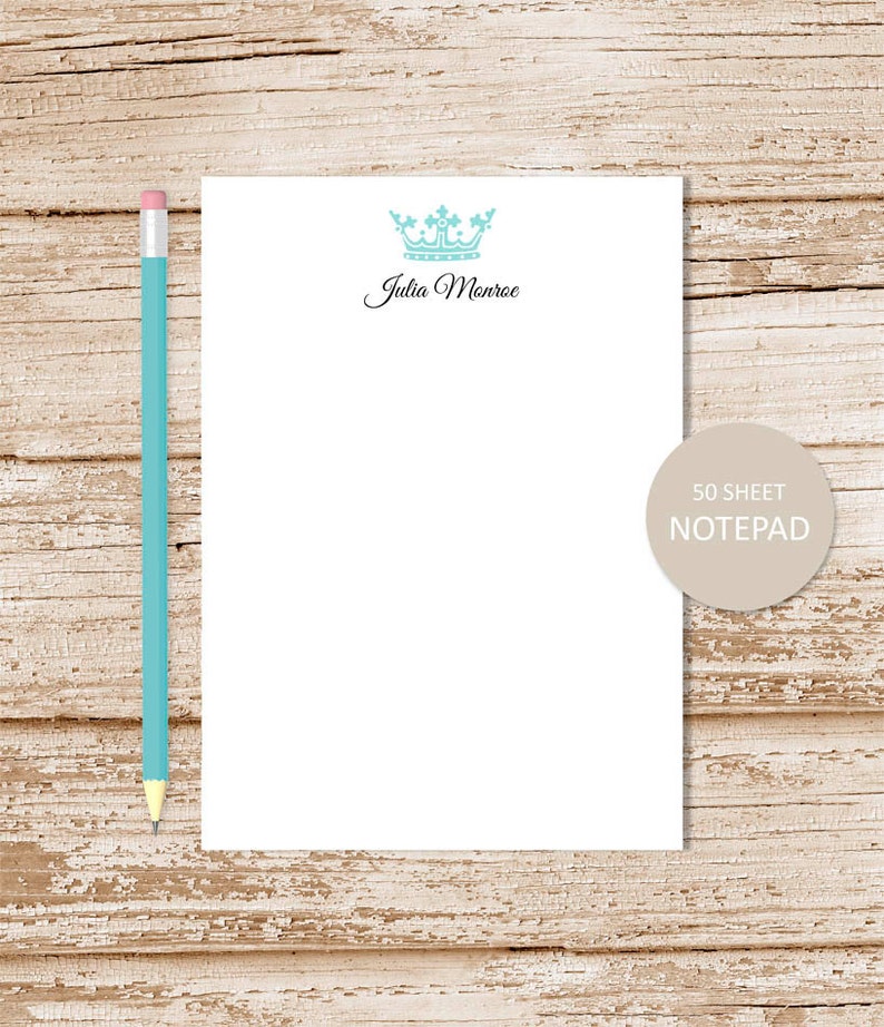 May include: A white notepad with a blue crown and the text "Julia Monroe" printed on the top. The notepad is on a wood background and has a label that says "50 sheet notepad".