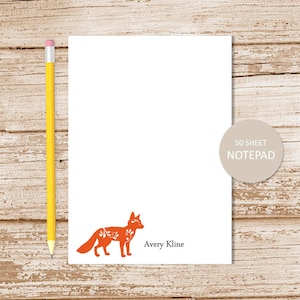 May include: White notepad with an orange fox illustration and the text "Avery Kline." A yellow pencil is positioned to the left of the notepad. The notepad has a circular label that reads "50 SHEET NOTEPAD."