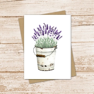 May include: Watercolor illustration of lavender in a white bucket with a handle. The bucket has a rustic, distressed appearance. The lavender has purple flowers and green stems. The image is on a white card with a brown envelope.