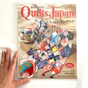 May include: A Quilts Japan magazine issue featuring a cover with a colorful patchwork quilt made of fabric petals, a small spherical fabric ball, and a stack of circular fabric swatches. The title text reads Quilts Japan in red letters.