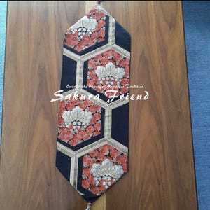 May include: A black and gold table runner with a geometric design. The runner features red and white floral patterns within hexagonal shapes. The text "Sakura Friend" and "Embrace the Beauty of Japanese Tradition" are visible.