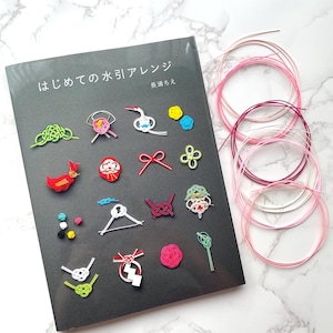 May include: A book titled "はじめての 水引アレンジ" with various colorful Mizuhiki craft examples. The book is on a dark gray surface, accompanied by several spools of pink and white Mizuhiki cord. The crafts include birds, flowers, and geometric shapes.