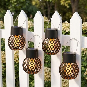 May include: Four solar-powered hanging lanterns with black metal lattice frames and cylindrical tops. Each lantern features a warm yellow light source inside the cage and a silver wire handle for suspension, displayed against a white picket fence.