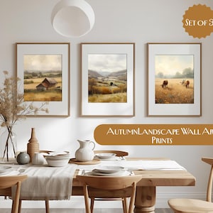 Op de afbeelding: Set van drie ingelijste landschapskunstprints. Elke print toont een ander herfsttafereel: een schuur in een veld, een vallei en koeien die grazen in een weide. De prints worden boven een eettafel weergegeven.