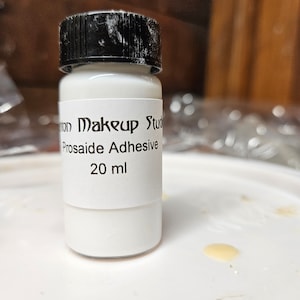 Puede incluir: Una botella blanca con tapa negra y etiqueta. La etiqueta dice "on Makeup Studio Prosaide Adhesive 20 ml".