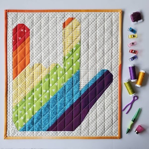 May include: A colorful quilted wall hanging in the shape of a hand sign. The hand is made of different fabric squares in shades of orange, yellow, green, blue, and purple. The background is white with black polka dots.