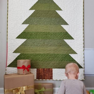 Pode incluir: Uma colcha de retalhos verde e marrom que representa uma árvore de Natal com uma estrela amarela no topo. Há presentes embrulhados em marrom com fitas vermelhas e douradas sob a árvore. Uma criança pequena está sentada no chão olhando para a colcha.