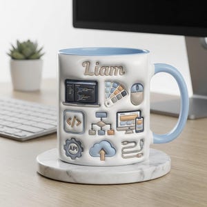 May include: White ceramic mug with a light blue handle and interior. The mug features the name "Liam" in a brown font, along with various computer-related icons. The mug is sitting on a marble coaster on a wooden desk.