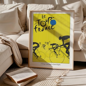 Puede incluir: Un póster enmarcado con fondo amarillo con las palabras "Le Tour de France" en negro y azul. Se representan ciclistas silueteados en bicicletas, con el texto "A long road and a hard fight" en la parte inferior. El póster está en un marco de madera clara.