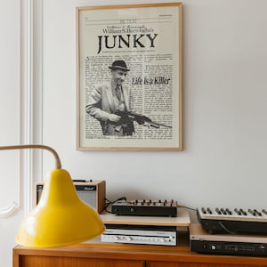 May include: A framed print of a newspaper page titled "Junky" by William S. Burroughs, featuring a black and white photo of a man holding a gun. The print is in a light wood frame. A yellow lamp and electronic equipment are in the foreground.
