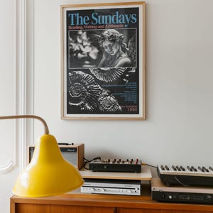 K&ouml;nnte beinhalten: Ein gerahmtes Poster mit dem Text "The Sundays" und "Reading, Writing and Arithmetic" &uuml;ber einem Schwarz-Wei&szlig;-Bild eines Engels und eines abstrakten Designs. Das Poster h&auml;ngt &uuml;ber einem Holzschrank mit einer gelben Lampe, einem Verst&auml;rker und Audioger&auml;ten.