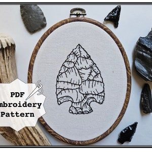 May include: A black and white embroidery pattern of an arrowhead in a wooden embroidery hoop. The hoop is surrounded by various arrowheads and a piece of antler.