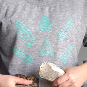 May include: A gray sweatshirt featuring a design of six turquoise-colored arrowheads. The arrowheads are outlined with a detailed, cracked pattern. The person is holding a piece of driftwood and a rock.