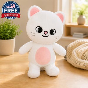 May include: A white plush cat toy with large black eyes, a small pink nose, and a light pink circular patch on its belly. The cat has rounded ears with pink inner details and embroidered whiskers on its cheeks, standing in an upright position.