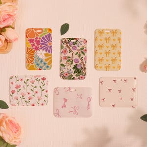 May include: A collection of six rectangular phone cases with various floral and bow designs. The cases feature vibrant colors and patterns, including orange flowers, purple daisies, and fig branches. Some cases have a bow pattern.