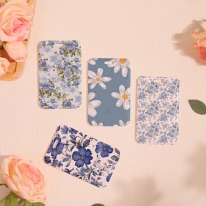 May include: Four phone cases with floral patterns. The cases feature blue and white floral designs, including roses and daisies, on a light background. One case has a blue background with white daisies. The cases are rectangular with a hole at the top.
