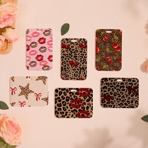 May include: Six rectangular luggage tags with diverse patterns. Designs include pink lips, leopard print, cherries, stars, and bows. Each tag has a small hole at the top for attachment.