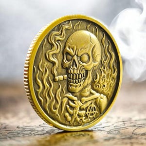 May include: A gold-colored coin featuring a detailed skull smoking a cigar, surrounded by flames. The coin has an anarchist symbol and is set against a blurred background, with smoke rising.